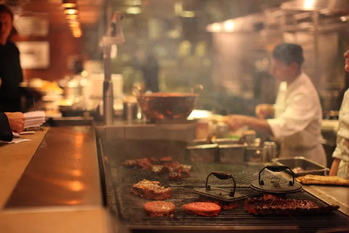 Hamburguesas en una parrilla y cocineros preparando pedidos al fondo.